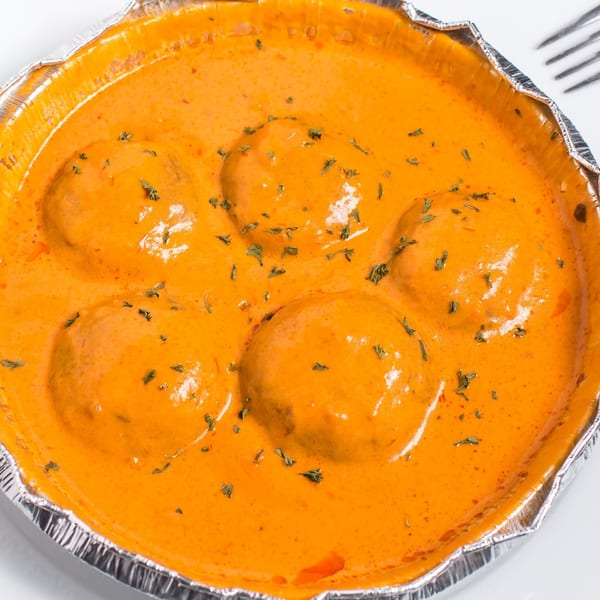 Five kofta balls in a rich orange curry sauce, garnished with green herbs, in a foil container.